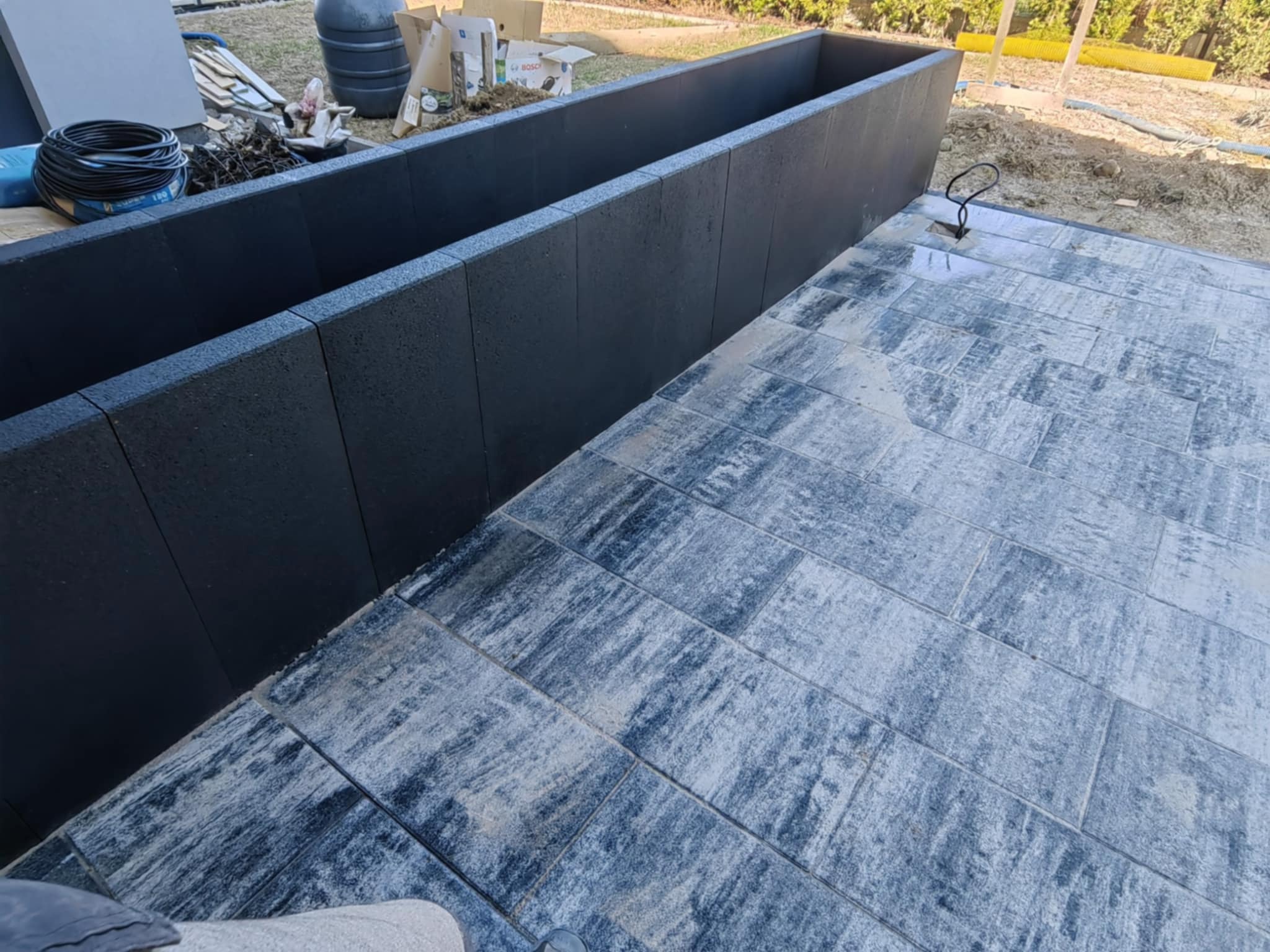 Nowoczesny taras z szaro-niebieskiej kostki, obok podwyższona rabata z czarnych bloków. Widoczne narzędzia i materiały budowlane. Ujęcie z góry.