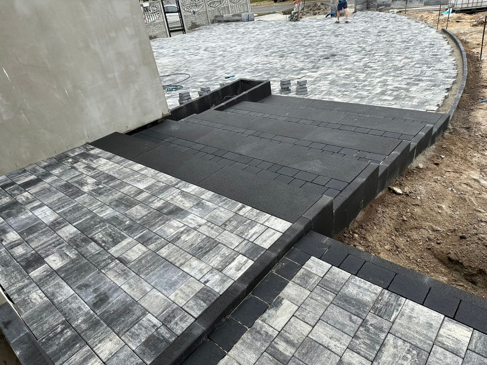 Kaskada z kostki brukowej w odcieniach szarości i czerni, prowadząca do domu. Częściowo ułożona nawierzchnia z widocznymi etapami prac.