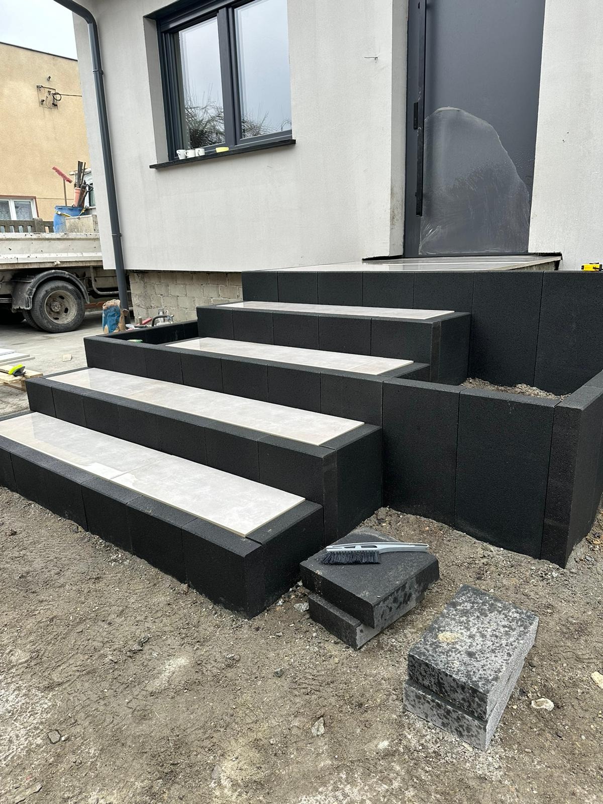 Nowoczesne schody wejściowe z czarnych bloków i jasnych płyt na wierzchu, prowadzące do domu z szarą elewacją i czarnymi drzwiami. Widoczne narzędzia brukarskie.