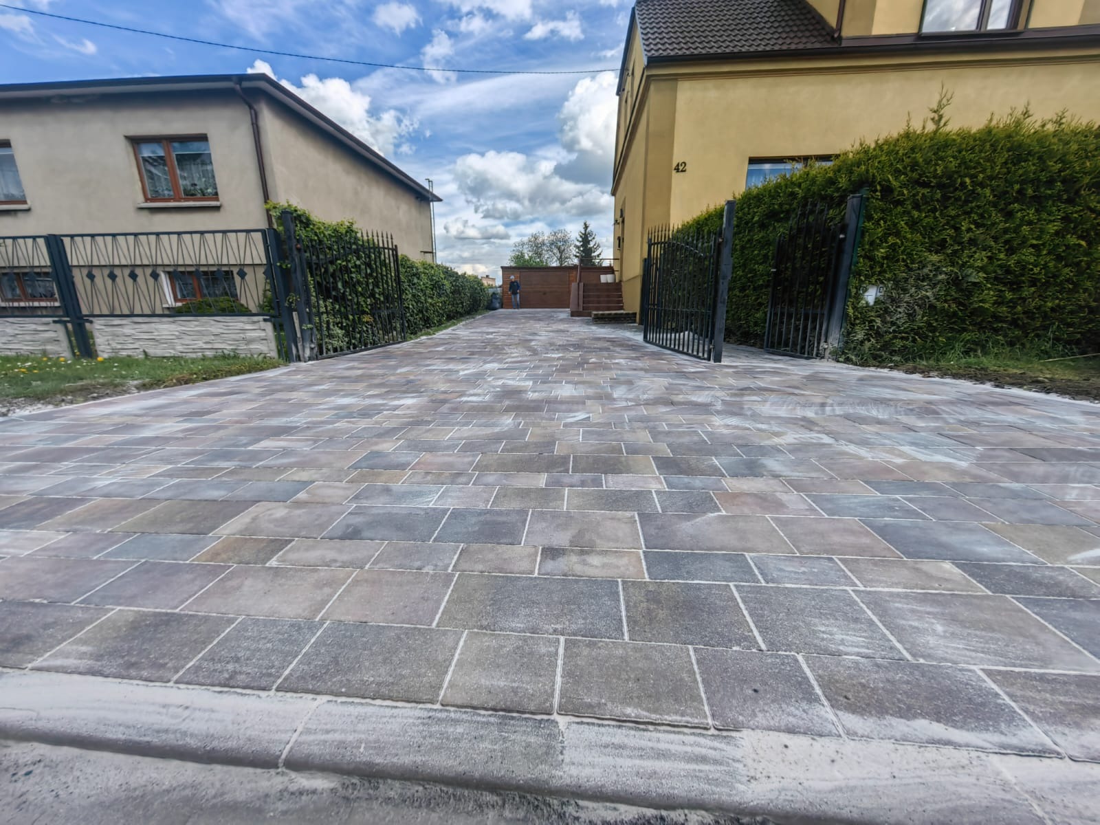 Szeroki, brukowany podjazd z kostki w odcieniach szarości i beżu, prowadzący do otwartej bramy i domu z numerem 42. Po bokach metalowe ogrodzenia i zieleń.
