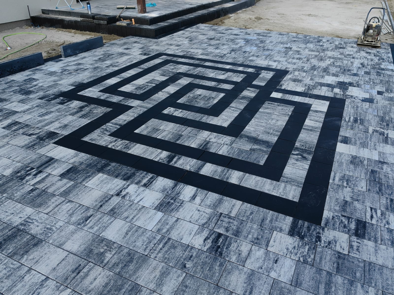 Ułożona kostka brukowa w odcieniach szarości z geometrycznym wzorem z czarnej kostki. W tle widoczny ubijak wibracyjny i fragment schodów.