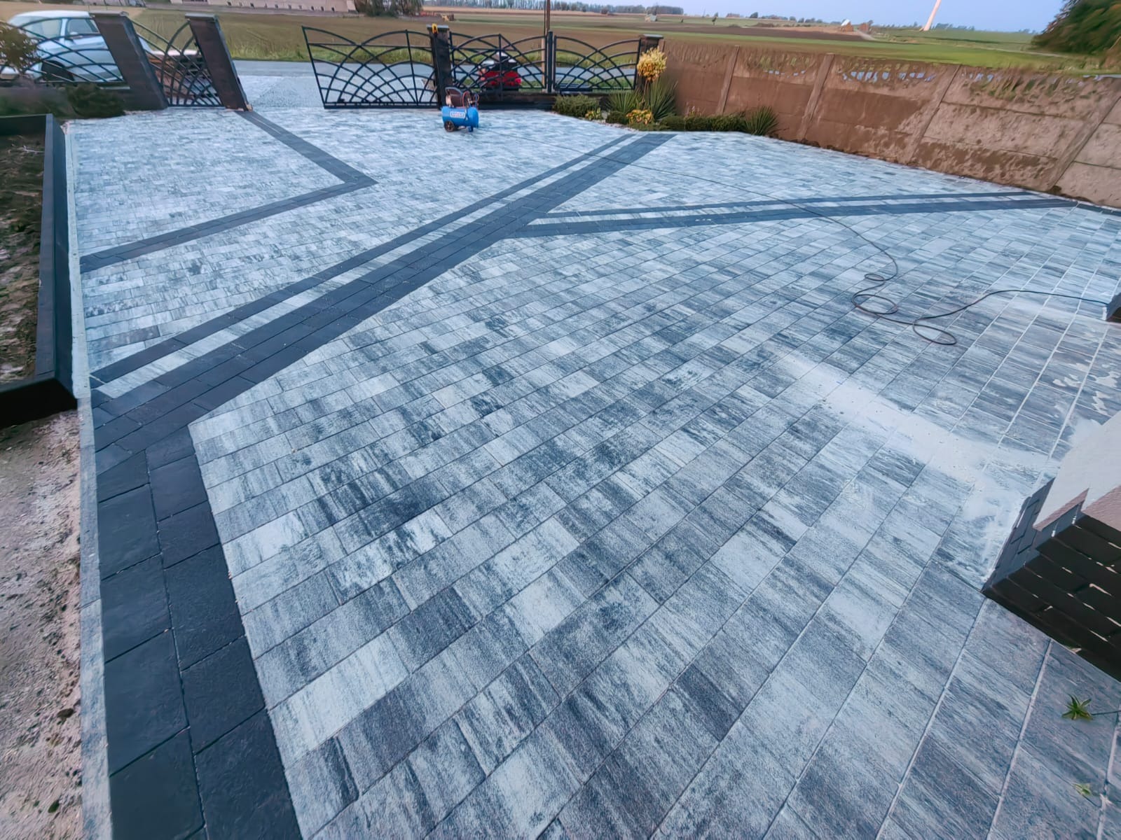 Ułożona kostka brukowa w odcieniach szarości z ciemnymi obramowaniami, tworząca wzór na podjeździe. Widoczna brama w tle i fragment ogrodu.