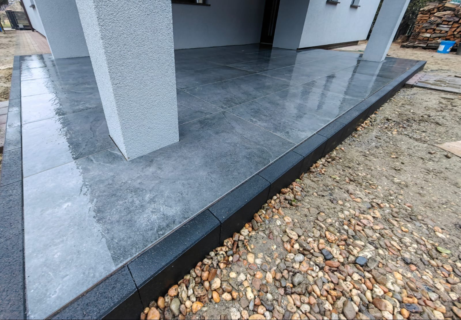 Nowoczesny taras z szarych płytek, obrzeża z czarnej kostki. Widoczne kamyki dekoracyjne. Minimalistyczny design, wysoka jakość wykonania.