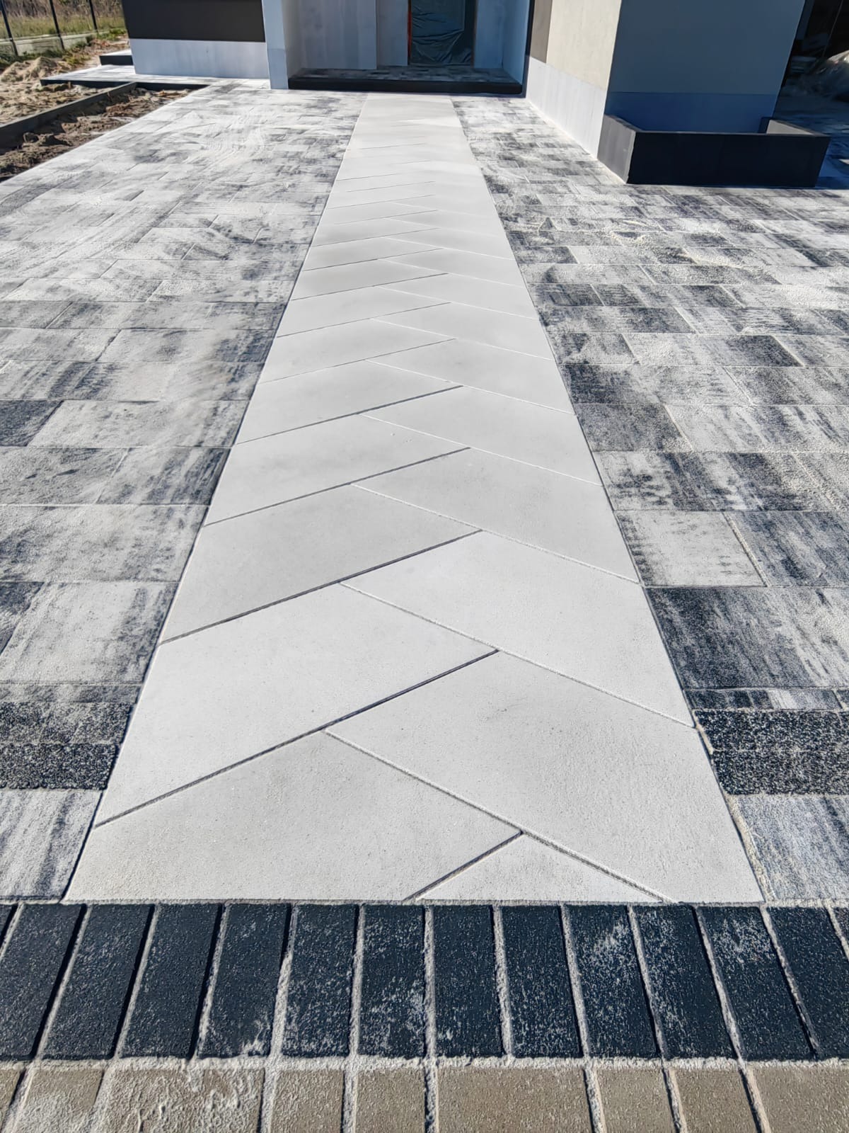 Ułożona kostka brukowa w odcieniach szarości prowadząca do budynku. Centralna ścieżka z jasnych, prostokątnych płyt ułożonych w jodełkę.