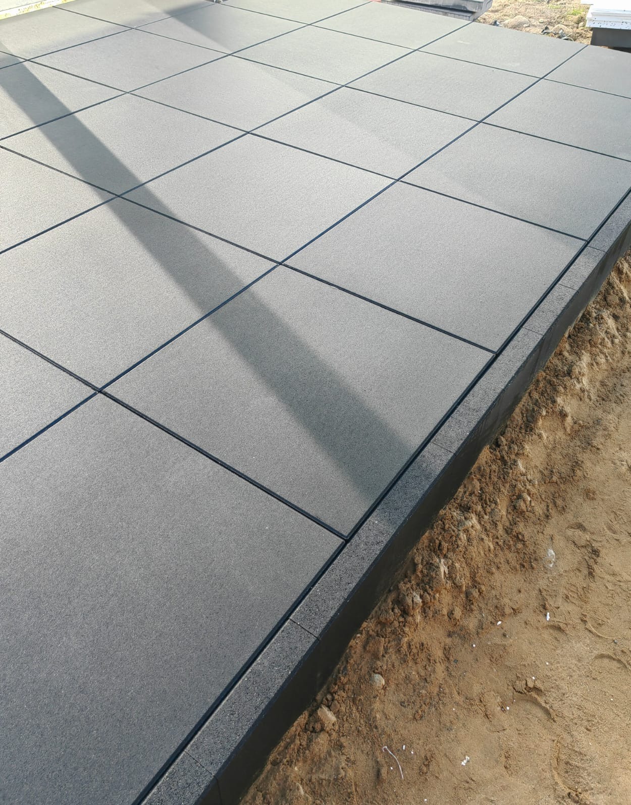 Nowoczesna, szara kostka brukowa ułożona w kwadraty z wąskimi fugami. Widoczny obrzeże i fragment podłoża z piasku. Cień na powierzchni kostki.