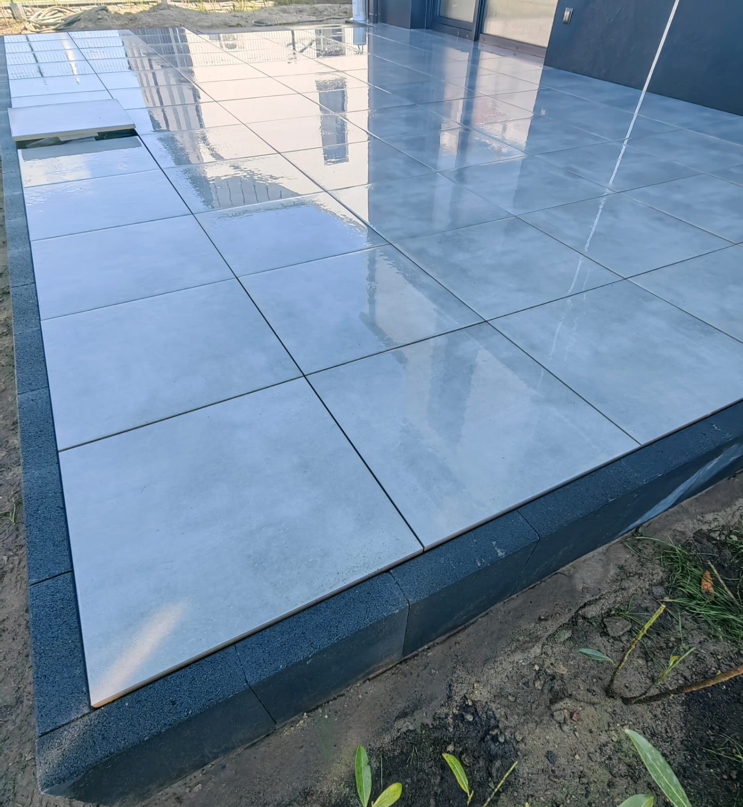 Jasnoszary taras z płytek w trakcie układania, z widoczną czarną obwódką. Kilka płytek jeszcze nie położonych. Odbicie budynku w błyszczącej powierzchni.