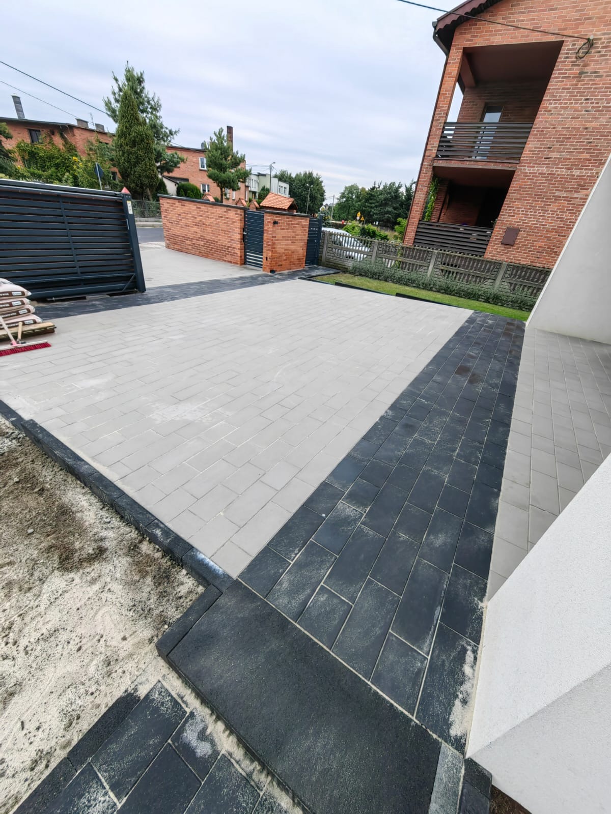 Jasnoszara kostka brukowa z ciemnymi obrzeżami tworzy wzór na podjeździe. W tle brama i fragment domu z czerwonej cegły. Widoczne ślady piasku.