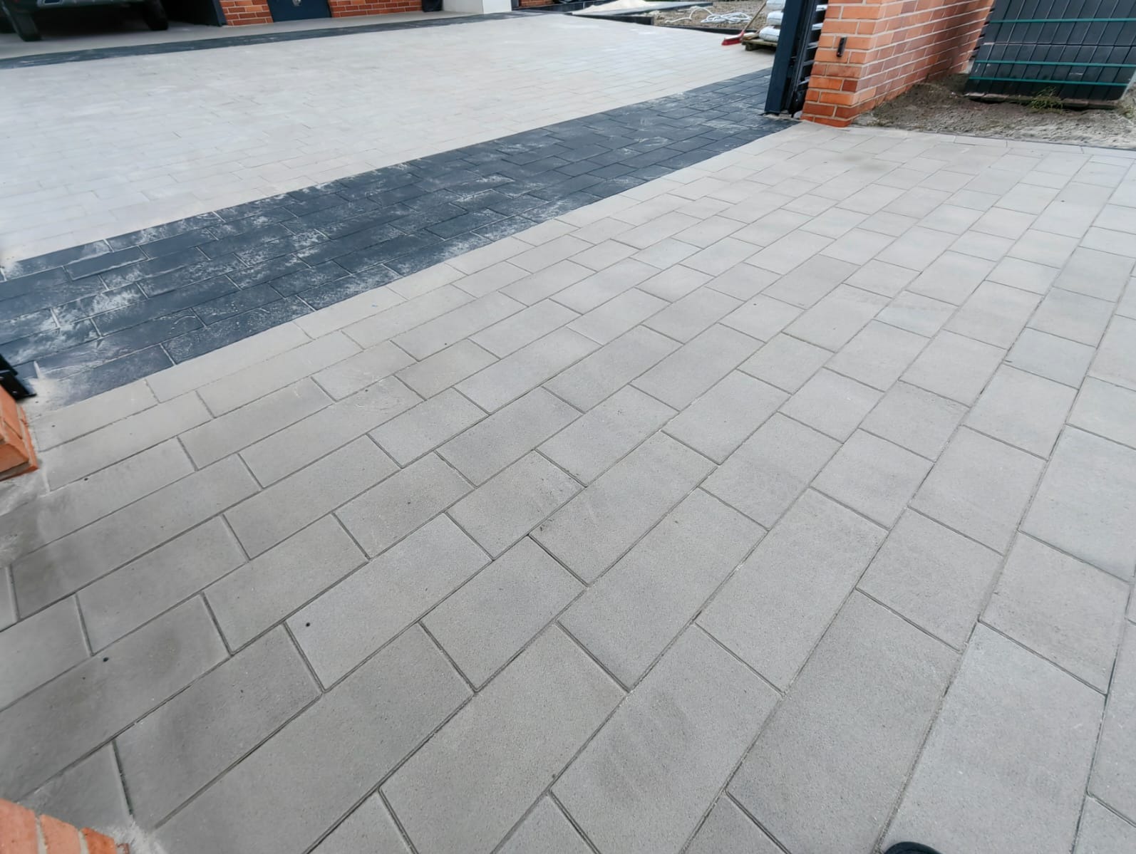 Nowoczesna nawierzchnia z prostokątnej kostki brukowej w odcieniach szarości, z ciemnym pasem pośrodku, prowadząca do bramy z ceglanym murem.