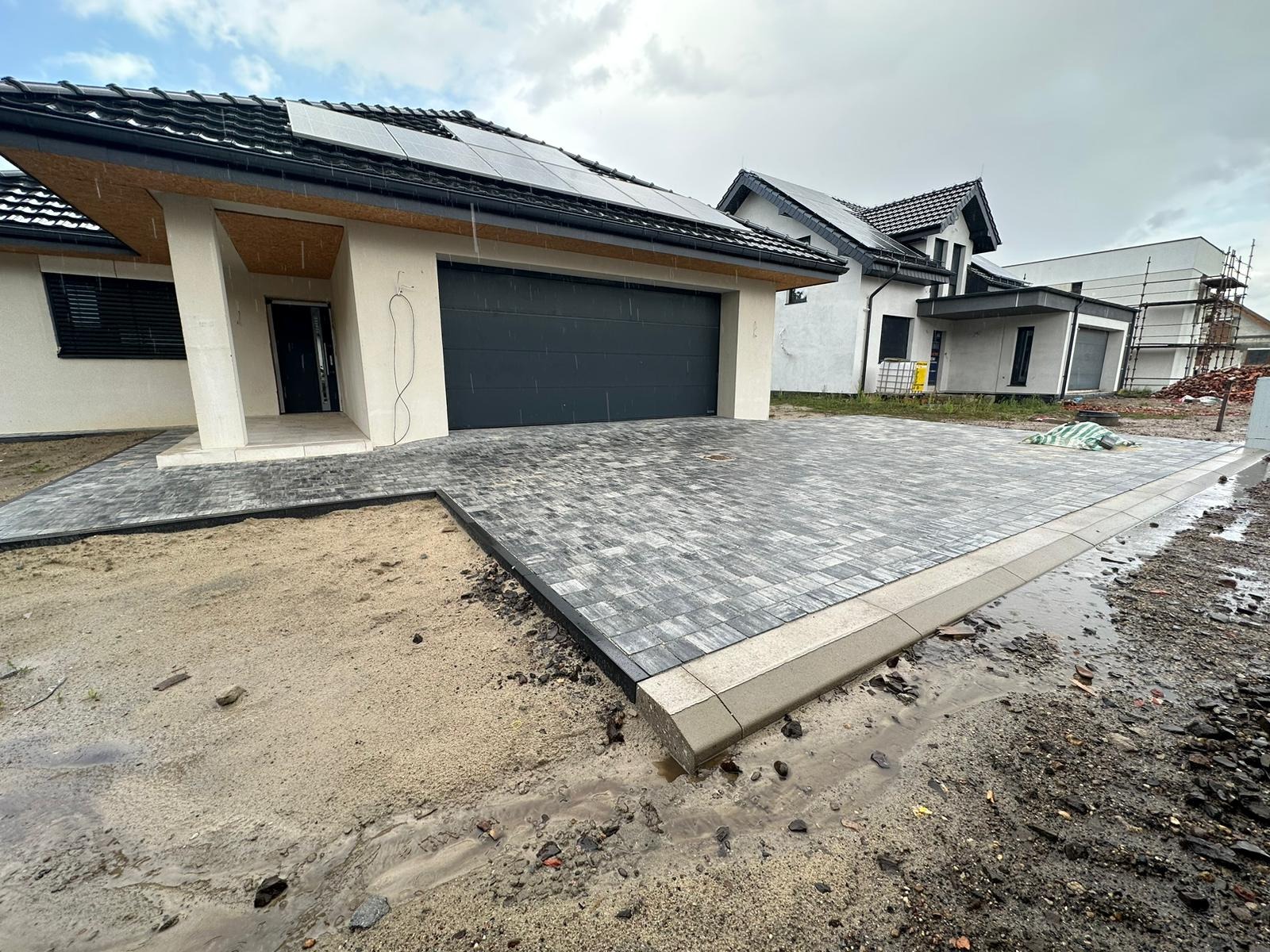Ułożona kostka brukowa z obrzeżami na podjeździe przed nowoczesnym domem z garażem i panelami słonecznymi. Częściowo podmokły teren.