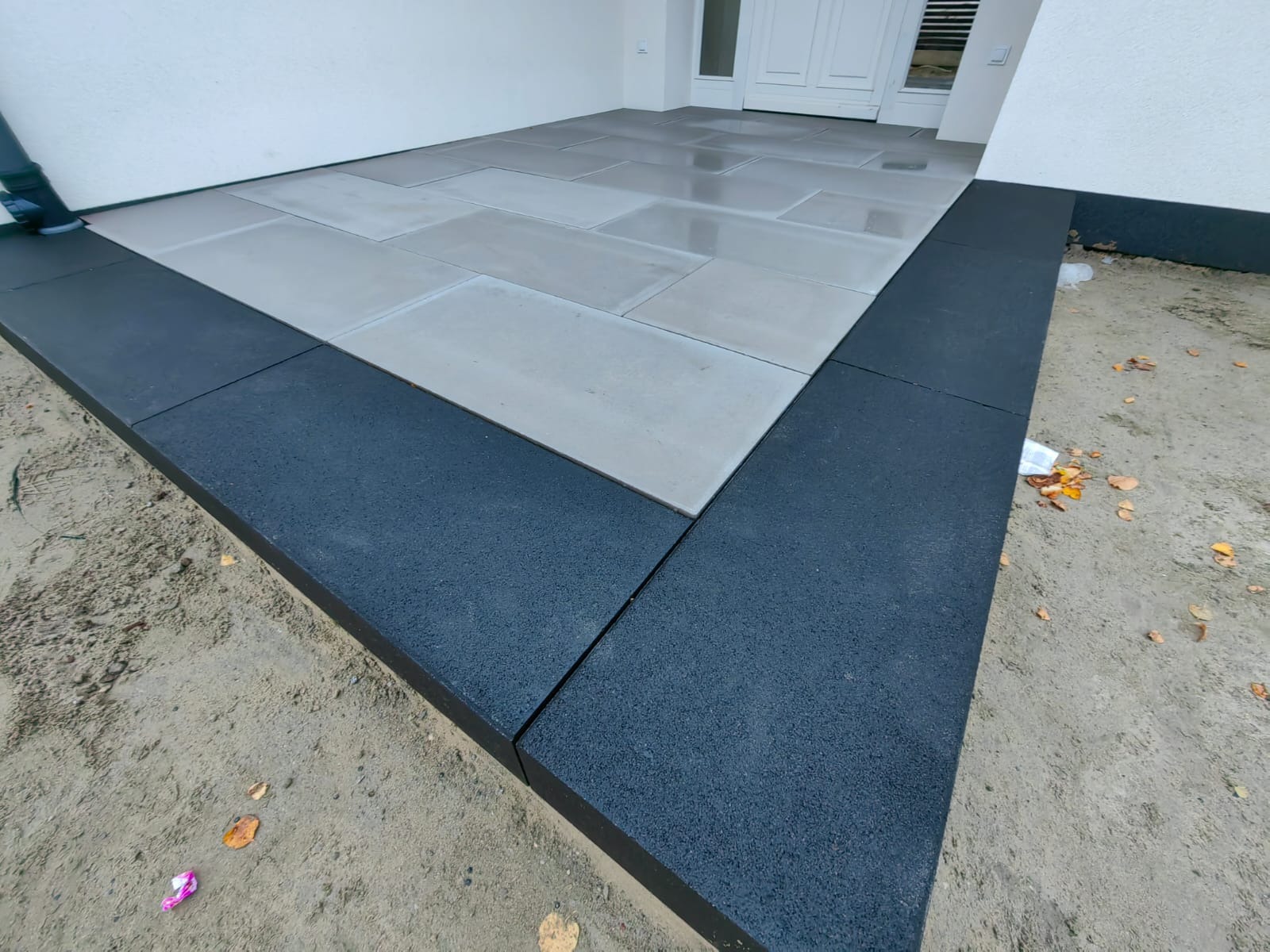 Nowoczesne wejście do domu z jasnoszarej kostki brukowej, obramowane ciemnoszarą kostką. Widoczne detale łączeń i faktura materiałów, podkreślające precyzję wykonania.