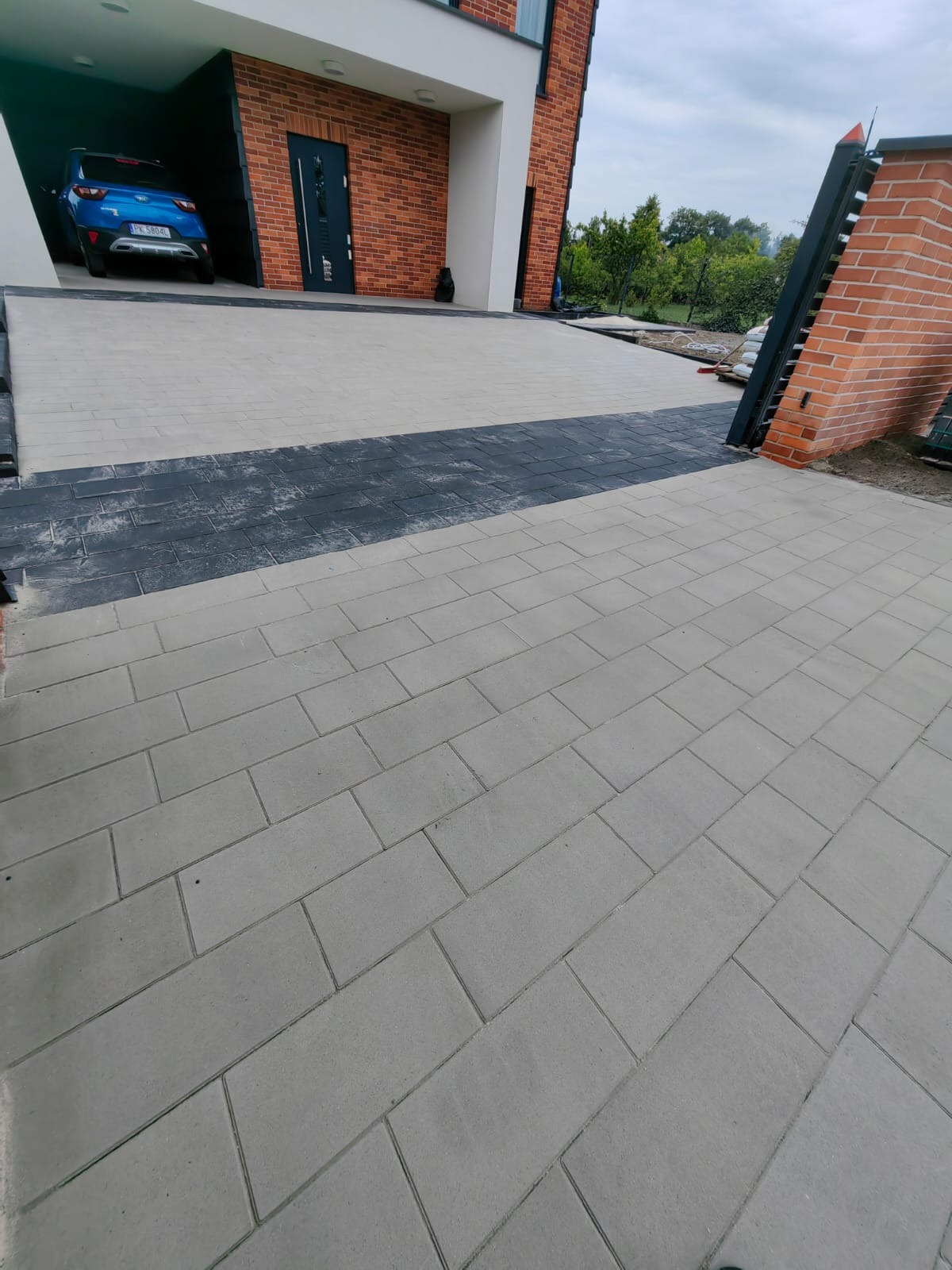 Ułożona kostka brukowa na podjeździe do garażu z widoczną bramą wjazdową z ceglanym słupkiem. Ciekawa kompozycja kolorystyczna kostki brukowej.