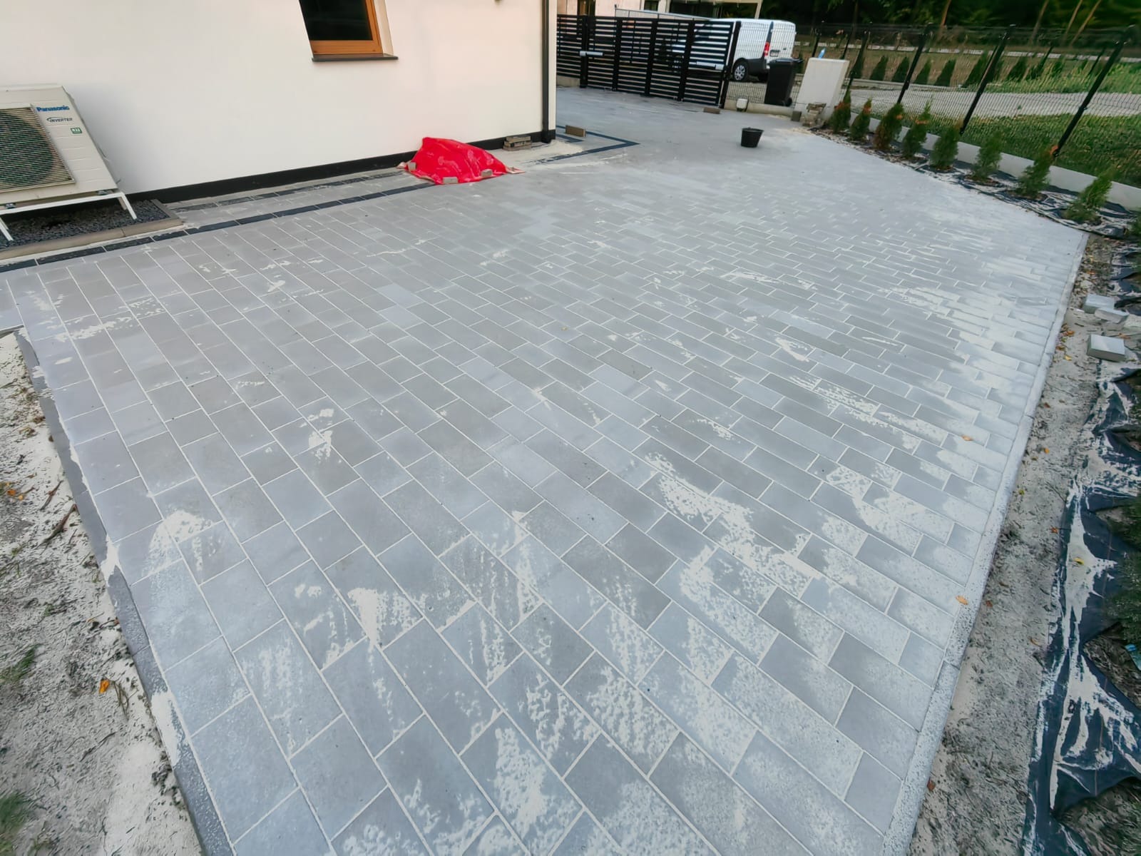 Świeżo ułożona, szara kostka brukowa w Biadkach, tworząca równy i estetyczny podjazd. Widoczne obrzeża, trawnik i fragment domu z klimatyzatorem.