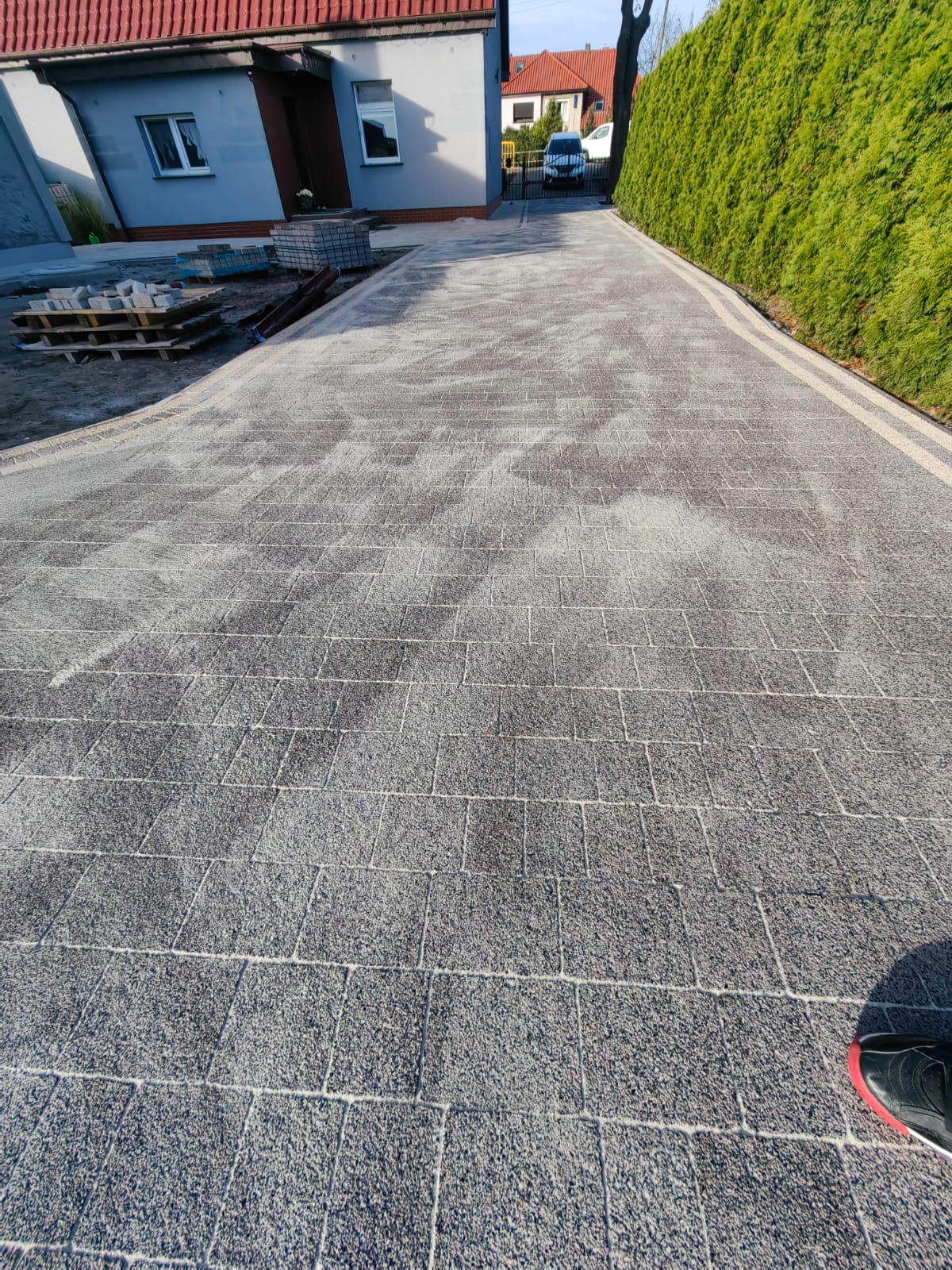Szara kostka brukowa ułożona na podjeździe, z widocznym fragmentem domu i zielonym żywopłotem. Ułożona kostka prowadzi w kierunku domu.
