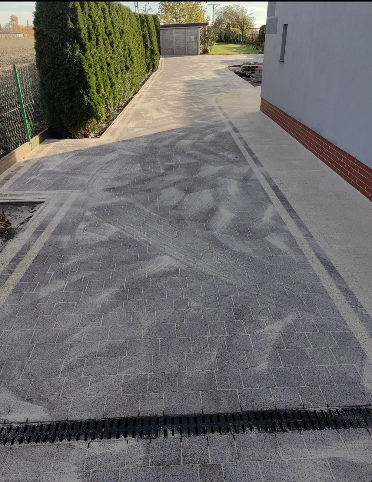 Szara kostka brukowa ułożona wzdłuż budynku i żywopłotu, z widocznym odpływem liniowym. Ślady piasku na powierzchni kostki, sugerujące świeżo zakończone prace.