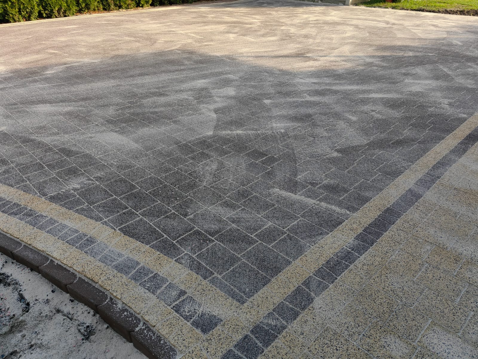 Szara kostka brukowa z dekoracyjnym obramowaniem w odcieniach beżu i brązu, posypana piaskiem. Widok z góry na dużą powierzchnię wyłożoną kostką.