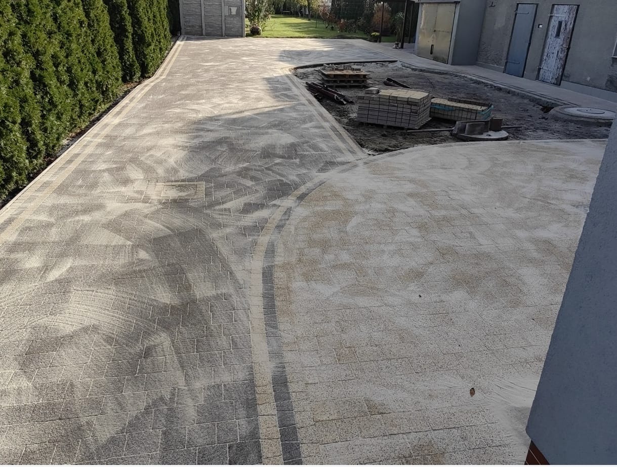 Ułożona kostka brukowa w odcieniach szarości tworzy ścieżkę z dekoracyjnymi obramowaniami. Część kostki jeszcze nieoczyszczona, w tle budynek gospodarczy i zieleń.