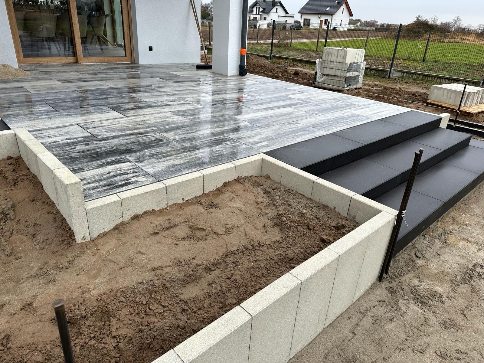 Nowoczesny taras z szaro-białej kostki, z czarnymi schodami i obrzeżem z jasnych bloczków. Częściowo ukończony, z widoczną ziemią w obrzeżu.