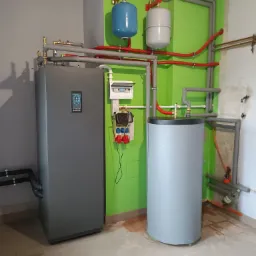 Szara instalacja grzewcza z zbiornikiem i licznikiem, rurami w izolacji, na tle zielonej i szarej ściany w pomieszczeniu.