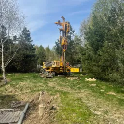 Żółta wiertnica do studni głębinowych na trawiastym terenie z widocznym operatorem i stertą rur w tle, otoczona drzewami.