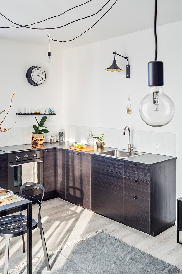 Nowoczesna kuchnia z ciemnymi, drewnopodobnymi szafkami, minimalistycznym zlewem i baterią, oraz dekoracyjnymi żarówkami zwisającymi z sufitu na tle białych ścian.