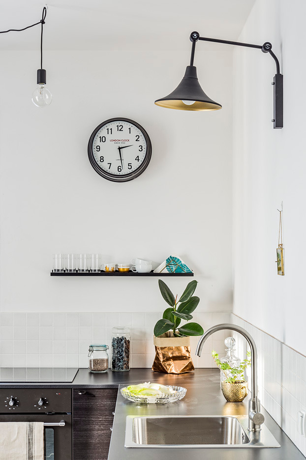 Nowoczesna kuchnia z metalowym blatem, minimalistycznym oświetleniem, zegarem London Clock i rośliną w złotej osłonce.