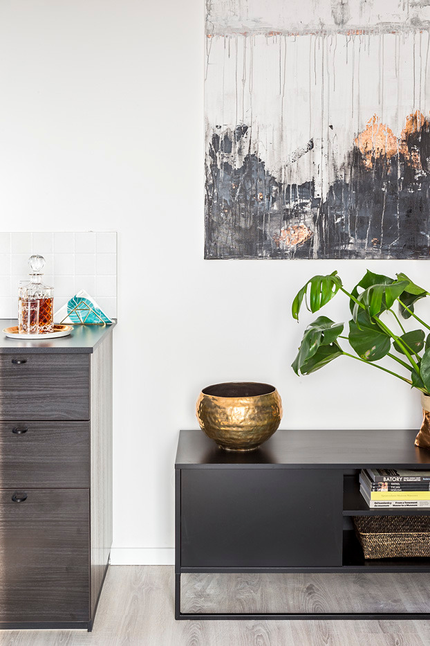 Nowoczesne wnętrze w Warszawie: ciemnoszara komoda z trzema szufladami, czarna szafka RTV z dekoracyjną, złotą misą i rośliną Monstera, abstrakcyjny obraz na ścianie, jasna podłoga.