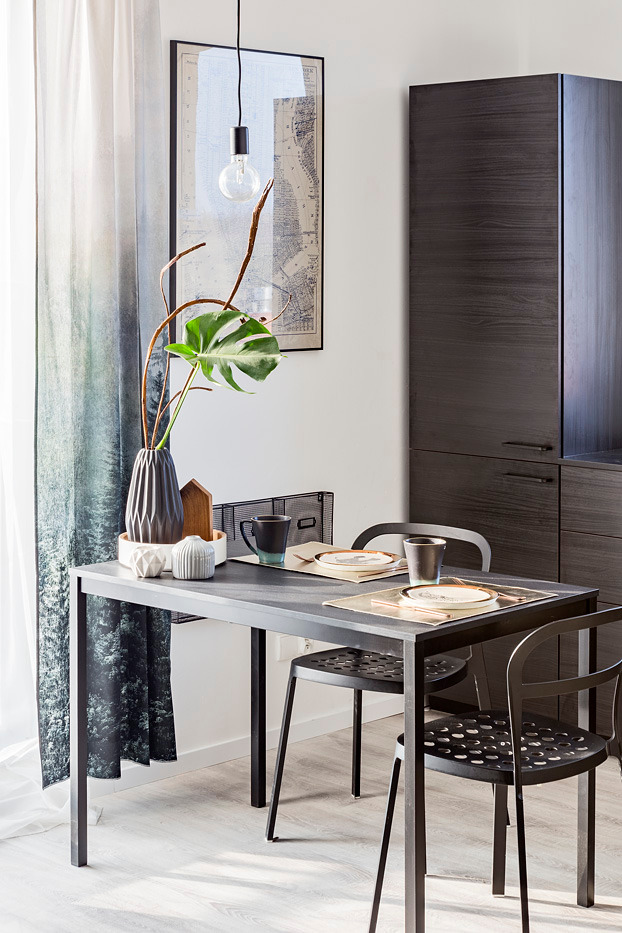 Nowoczesna jadalnia z czarnym stołem i krzesłami, minimalistyczna aranżacja z dekoracyjną rośliną monstera w wazonie, wisząca żarówka Edison i grafika na ścianie, obok ciemna zabudowa kuchenna.