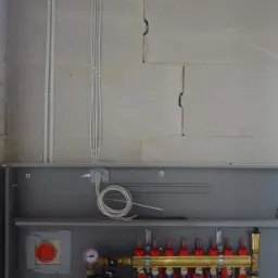 Rozdzielacz ogrzewania podłogowego w szarej szafce, z widocznymi rurami czerwonymi i niebieskimi, manometrami oraz przewodami elektrycznymi biegnącymi po ścianie z betonu komórkowego.