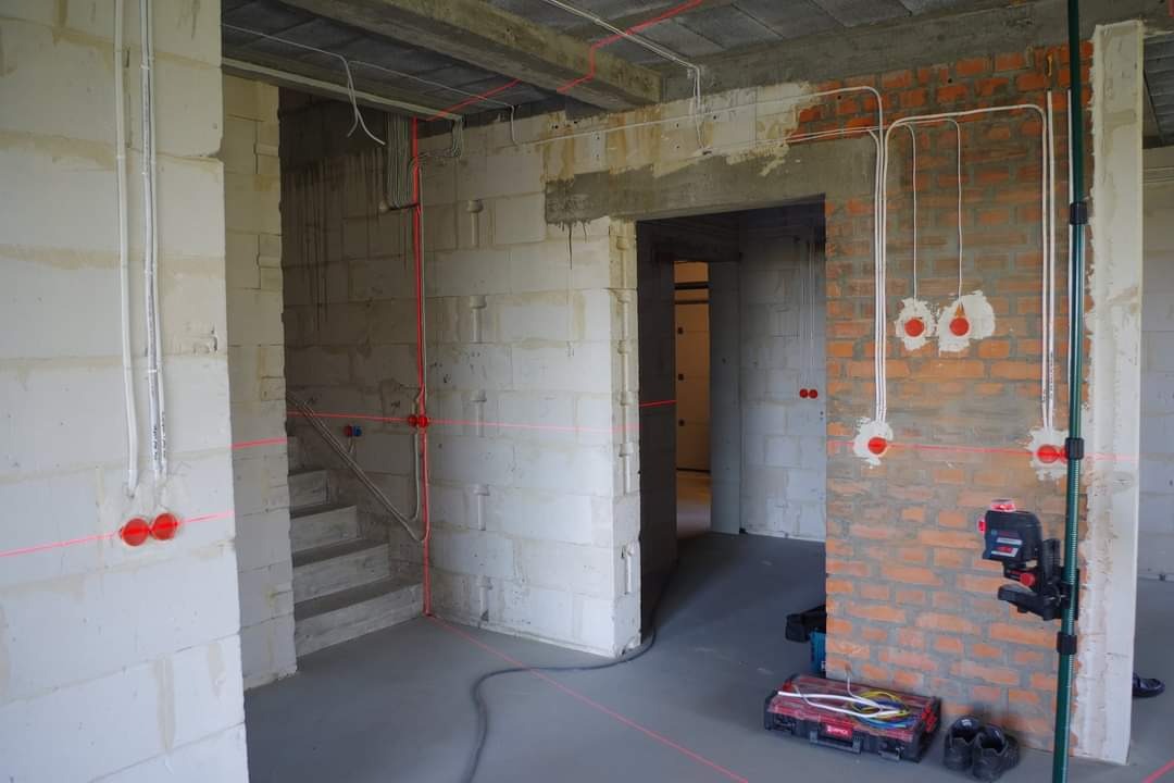 Wnętrze w trakcie prac wykończeniowych z widocznymi przewodami elektrycznymi, puszkami instalacyjnymi, poziomnicą laserową i narzędziami na podłodze.