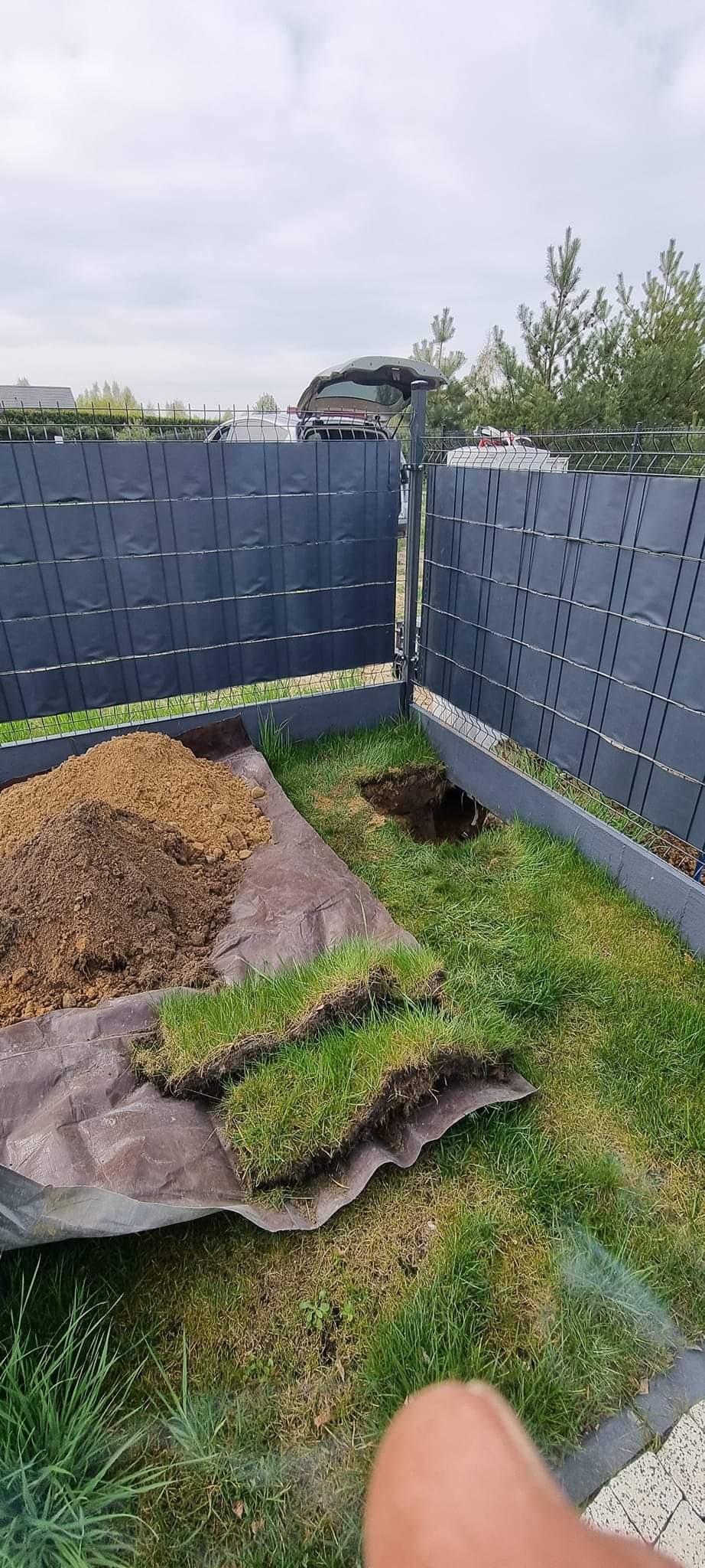 Wykop w trawniku przy ogrodzeniu panelowym, obok wycięte fragmenty darni i pryzma ziemi na plandece, w tle otwarty bagażnik samochodu.