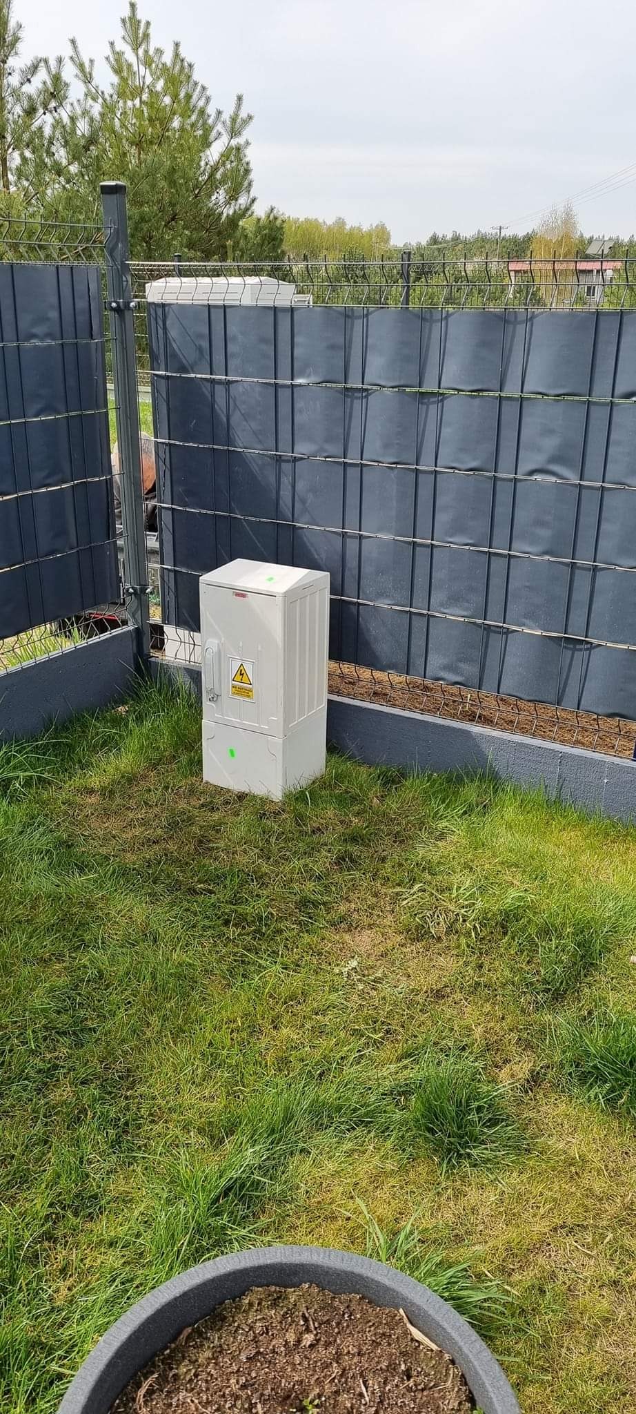 Biała skrzynka elektryczna z symbolem ostrzegawczym wysokiego napięcia, umieszczona na trawniku obok ogrodzenia panelowego.