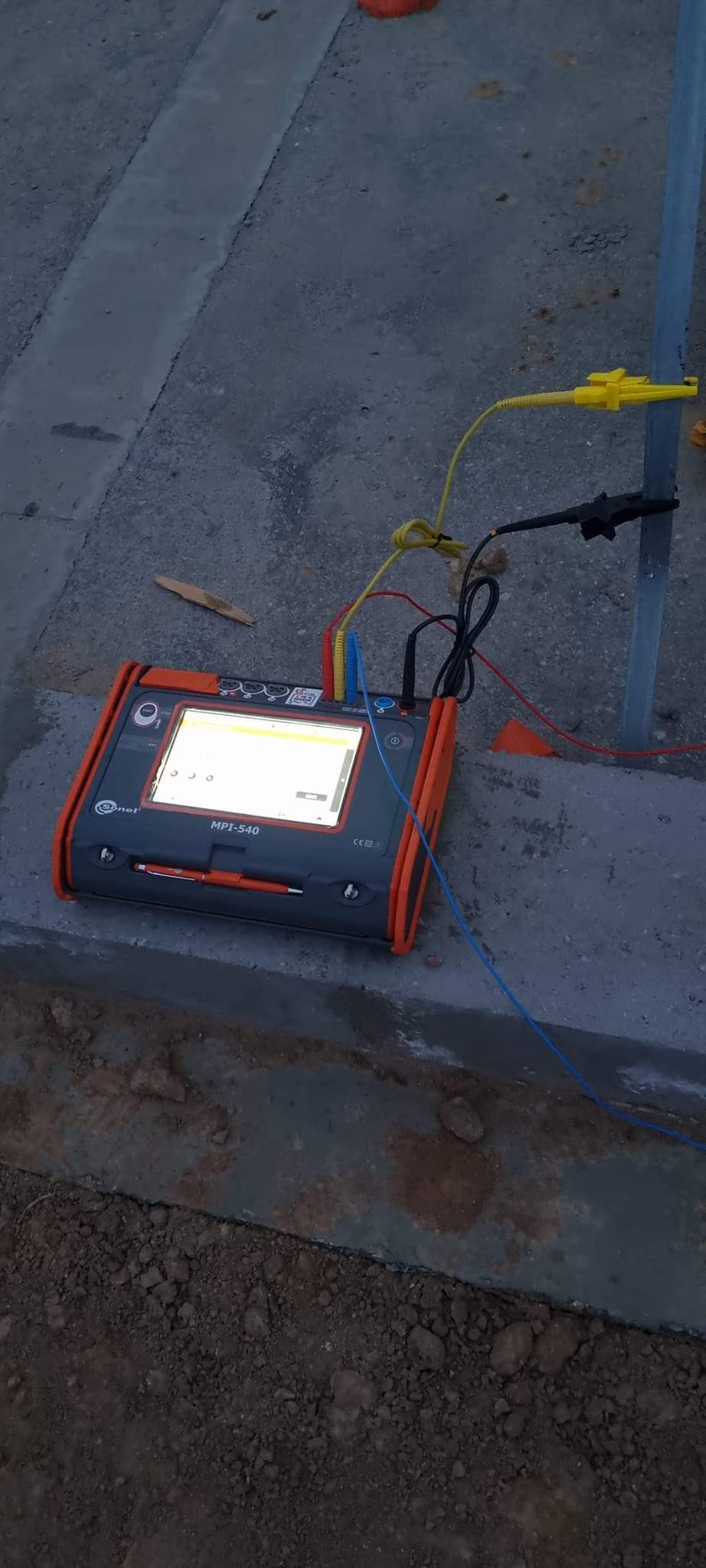 Miernik elektryczny Sonel MPI-540 z podłączonymi przewodami pomiarowymi, używany do testowania uziemienia metalowego słupka na zewnątrz budynku.