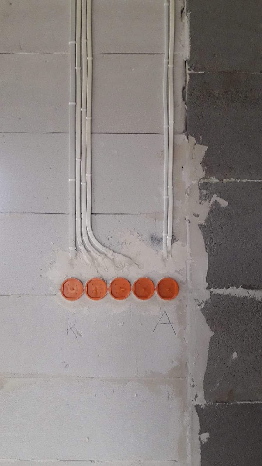 Białe kable elektryczne prowadzone wzdłuż ściany z betonu komórkowego, zakończone pomarańczowymi puszkami instalacyjnymi w rzędzie, widoczne ślady po ołówku na ścianie.