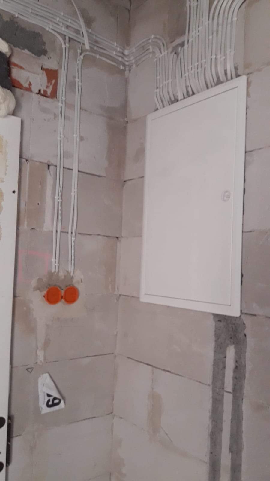 Biała skrzynka elektryczna zamontowana na surowej ścianie z betonu komórkowego, z widocznymi przewodami w białych peszlach ułożonymi w górnym rogu pomieszczenia i dwoma pomarańczowymi puszkami...