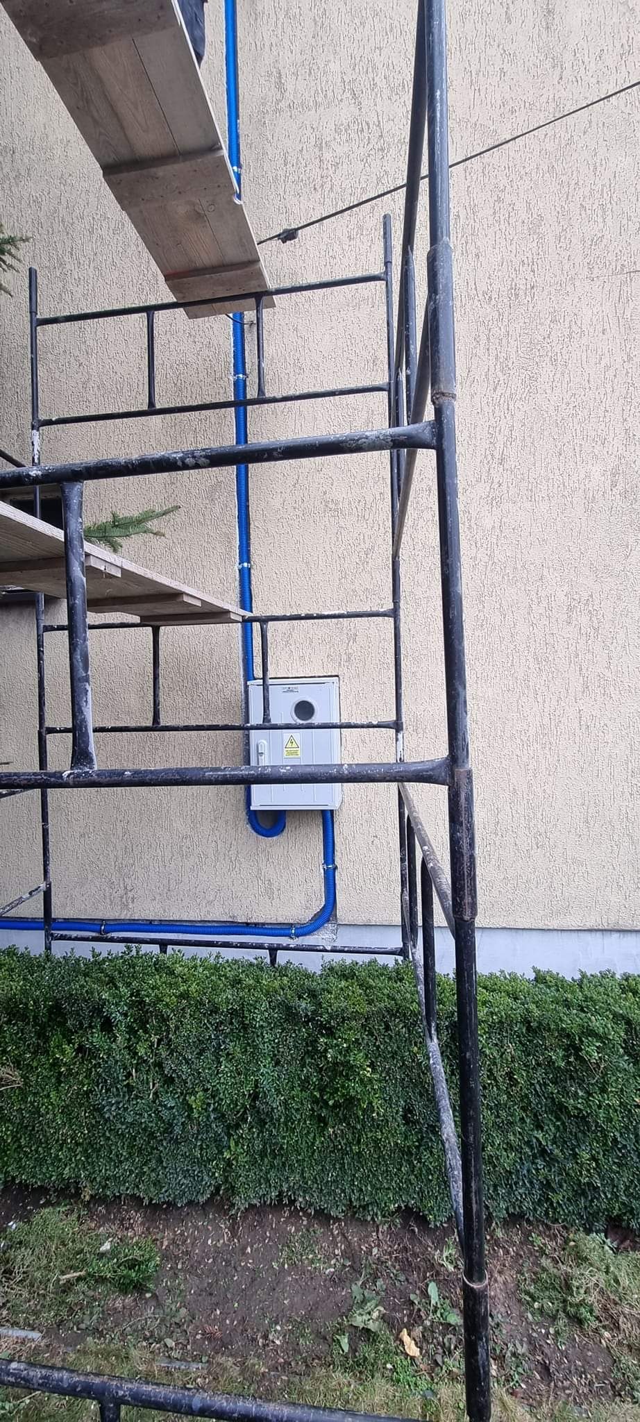 Montaż niebieskiej rury instalacji elektrycznej na elewacji budynku, widoczny fragment rusztowania i skrzynka elektryczna z symbolem ostrzegawczym.