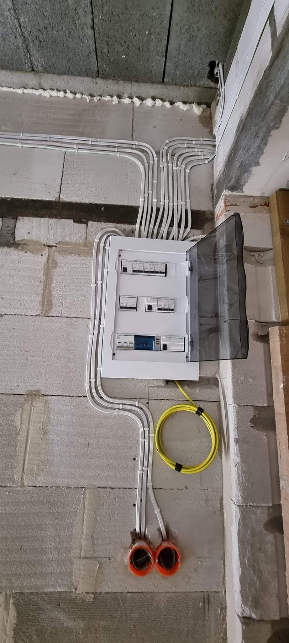 Rozdzielnica elektryczna z otwartą osłoną, widoczne liczne przewody w białych peszlach, żółty kabel zwinięty w pętlę i pomarańczowe puszki instalacyjne w surowej ścianie z betonu komórkowego.