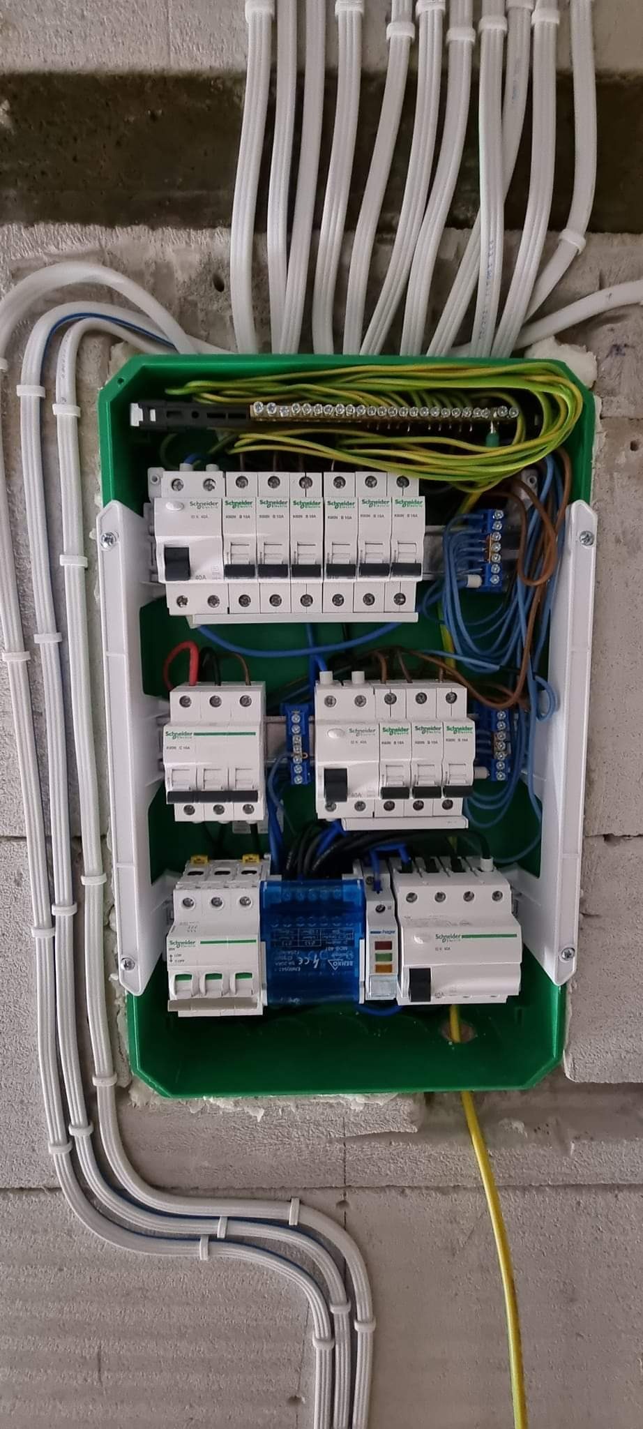 Zielona skrzynka rozdzielcza z zamontowanymi bezpiecznikami i przewodami Schneider Electric na surowej ścianie z betonu komórkowego, widoczne białe peszle z przewodami.