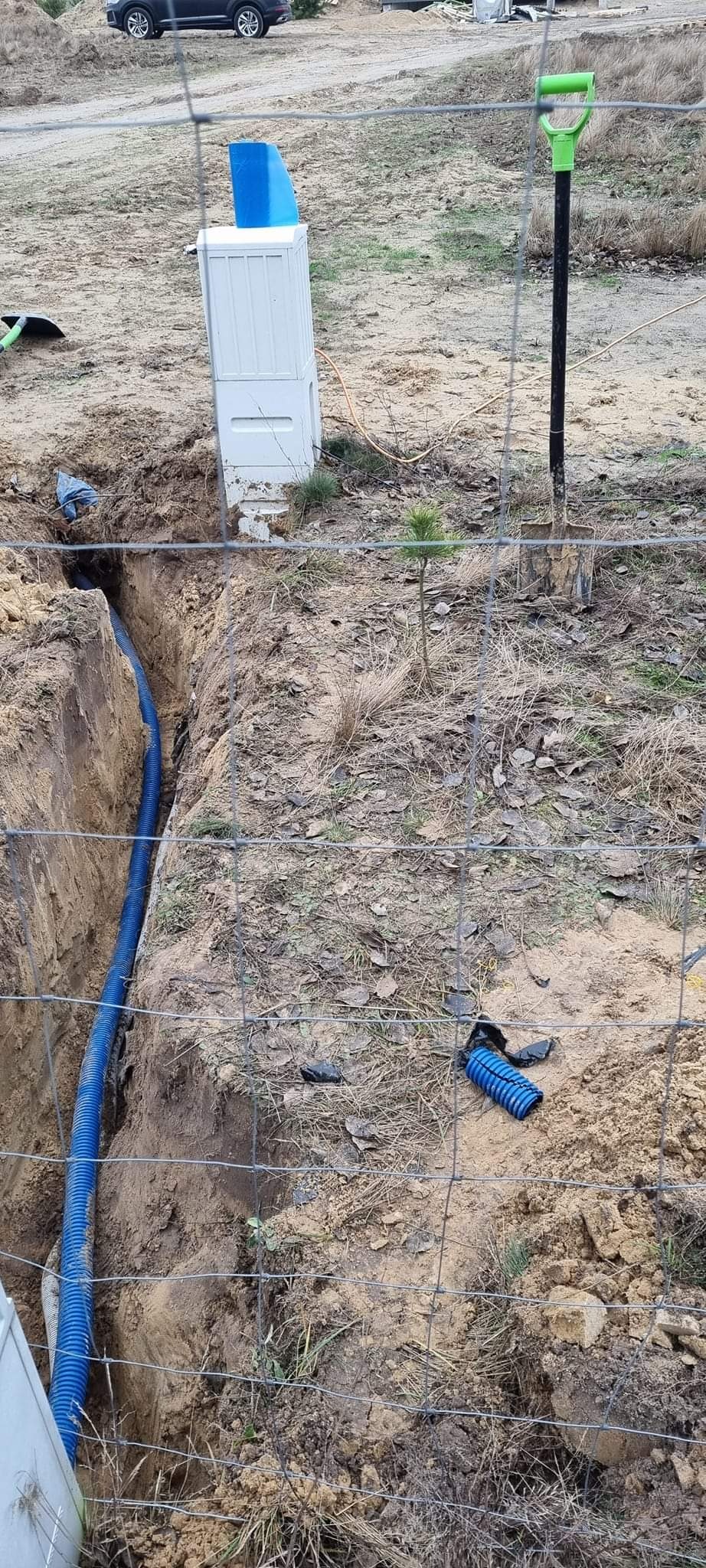 Niebieski przewód w wykopie, obok skrzynka elektryczna, widoczna łopata z zieloną rączką, teren ogrodzony siatką.