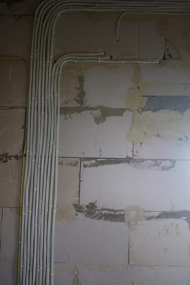 Białe kable elektryczne ułożone w peszlach, przymocowane do surowej ściany z betonu komórkowego. Widoczny fragment żółtego kabla. Ściana częściowo otynkowana, z widocznymi śladami po zaprawie.
