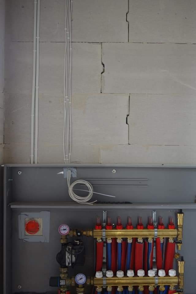 Rozdzielacz ogrzewania podłogowego w szarej szafce, z widocznymi rurami czerwonymi i niebieskimi, manometrami oraz przewodami elektrycznymi biegnącymi po ścianie z betonu komórkowego.