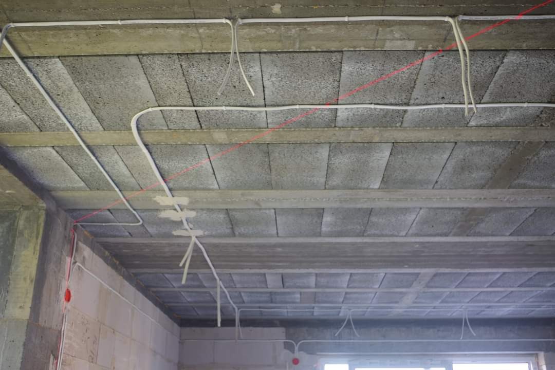 Instalacja elektryczna natynkowa z przewodami w peszlach prowadzona po betonowym stropie z pustaków szalunkowych, widoczna czerwona linia lasera wyznaczająca trasę.