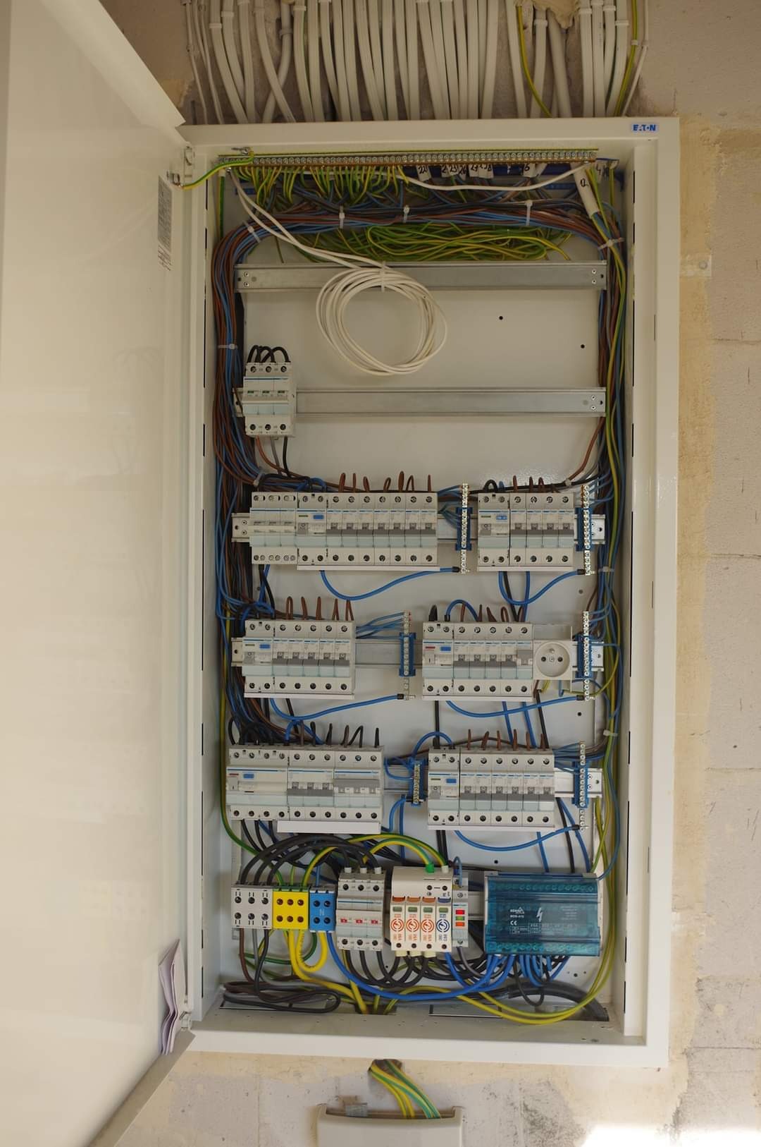 Otwarta skrzynka rozdzielcza z widocznymi bezpiecznikami, przewodami w różnych kolorach oraz listwami zaciskowymi, zamontowana na surowej ścianie z betonu komórkowego.