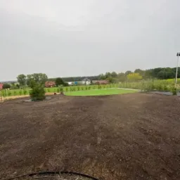 Przygotowany teren pod trawnik z rolki, widoczna świeżo rozplantowana ziemia i fragment ułożonej trawy w oddali na tle krajobrazu wiejskiego.