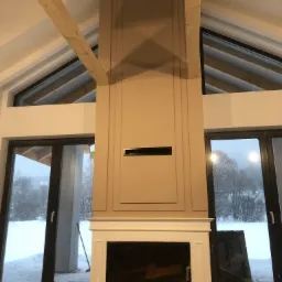 Wnętrze domu w trakcie wykończenia: beżowa obudowa kominka z dekoracyjnymi listwami, widok na zasypany śniegiem ogród przez duże okna, surowa drewniana belka stropowa