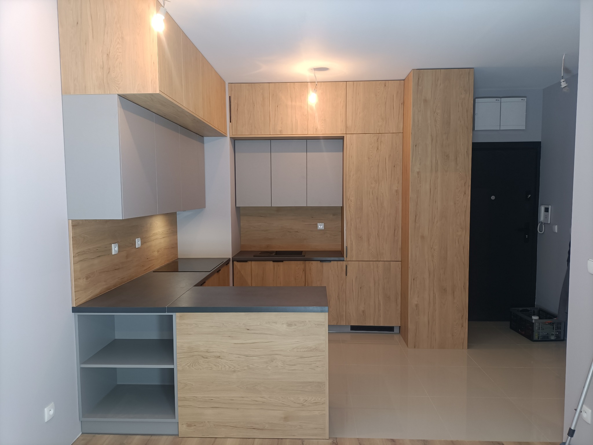 Nowoczesna kuchnia z jasnymi drewnianymi szafkami górnymi i szarymi dolnymi, ciemny blat i wyspa kuchenna z otwartymi półkami, widok na czarne drzwi w tle.