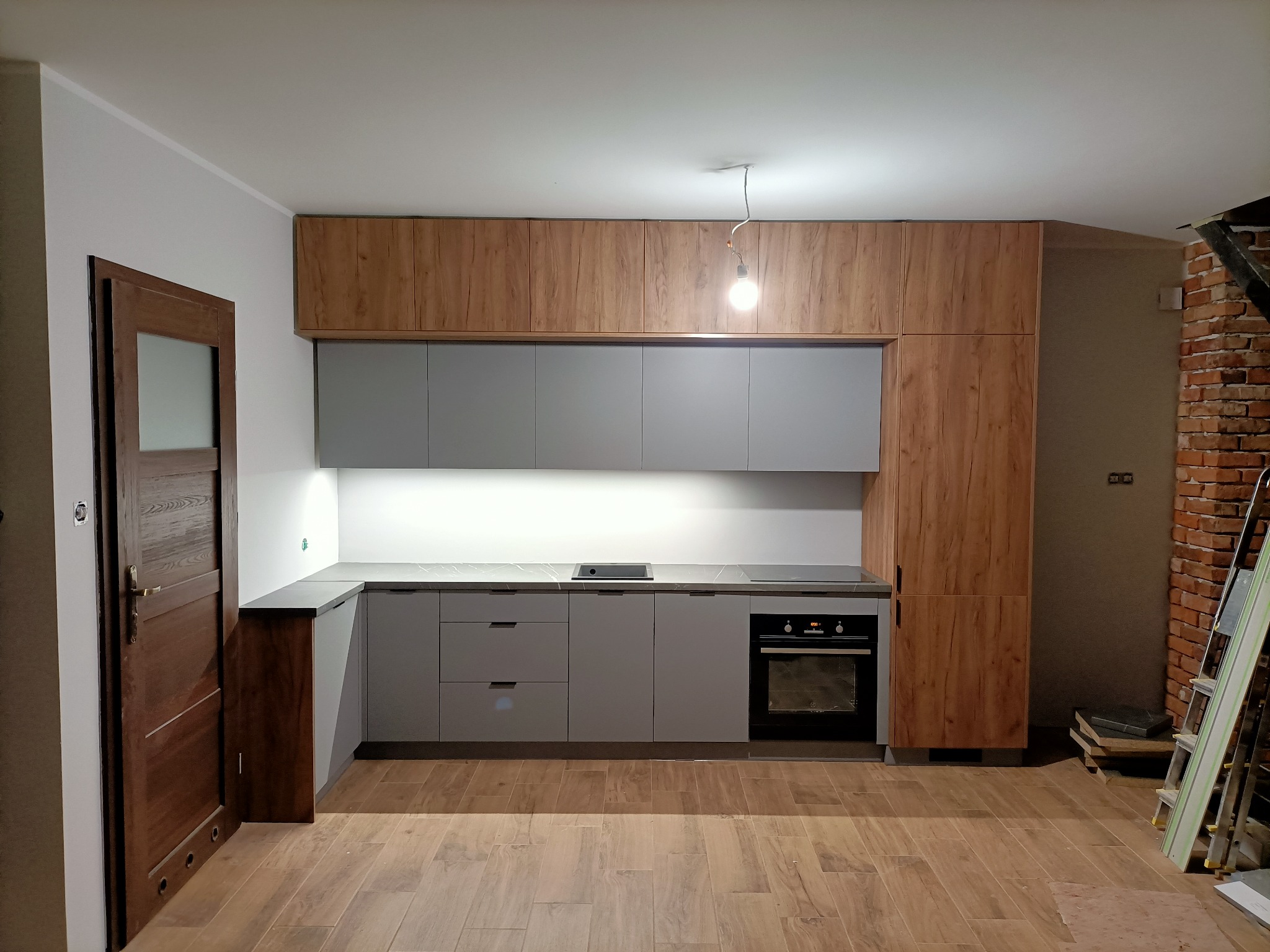 Nowoczesna kuchnia z szarymi frontami i drewnopodobnymi szafkami górnymi, widok na zabudowę kuchenną z piekarnikiem i płytą grzewczą, surowy ceglany mur w tle, podłoga wyłożona płytkami imitującymi...