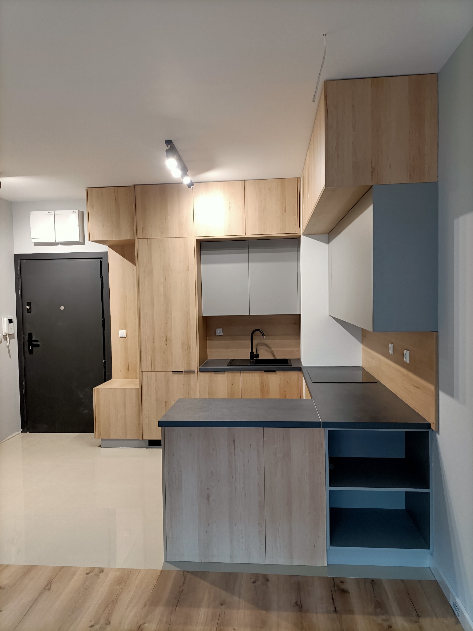 Nowoczesna kuchnia z drewnianymi frontami szafek i szarym blatem, czarny zlew i bateria, oświetlenie punktowe na suficie, widok na drzwi wejściowe