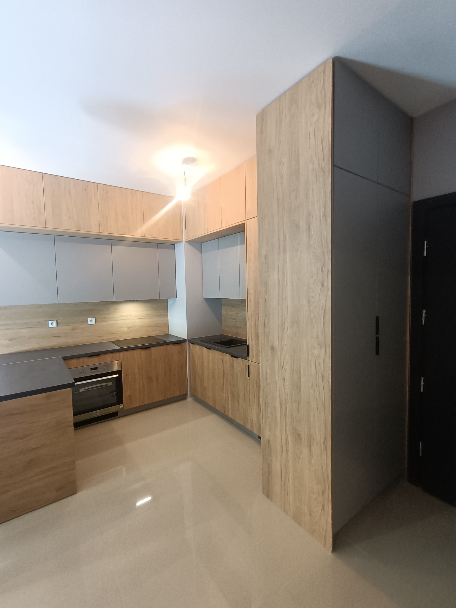 Nowoczesna kuchnia z drewnopodobnymi frontami szafek dolnych i szarymi szafkami górnymi, czarny blat i wysoka zabudowa z szarą szafą obok czarnych drzwi.