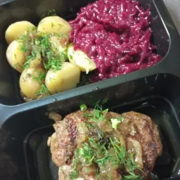 Gotowy posiłek w czarnym pojemniku: kotlet mielony z karmelizowaną cebulą i koperkiem obok gotowanych ziemniaków z sosem i posiekanej czerwonej kapusty.