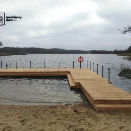 Drewniany pomost na jeziorze z drabinkami do wody i kołem ratunkowym, w tle zalesiony brzeg i pochmurne niebo.